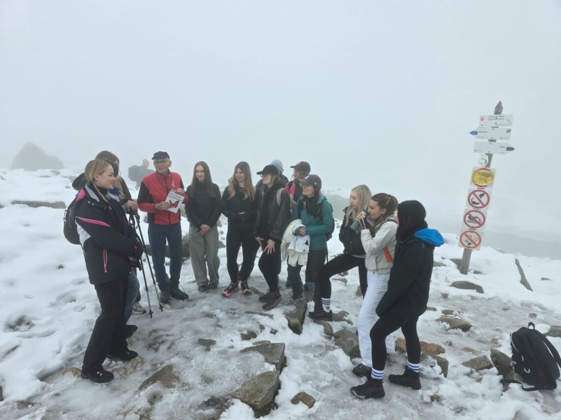 Grupa ludzi stojąca w półkolu na śnieżnym szlaku, z tyłu drogowskaz, widać mgłę